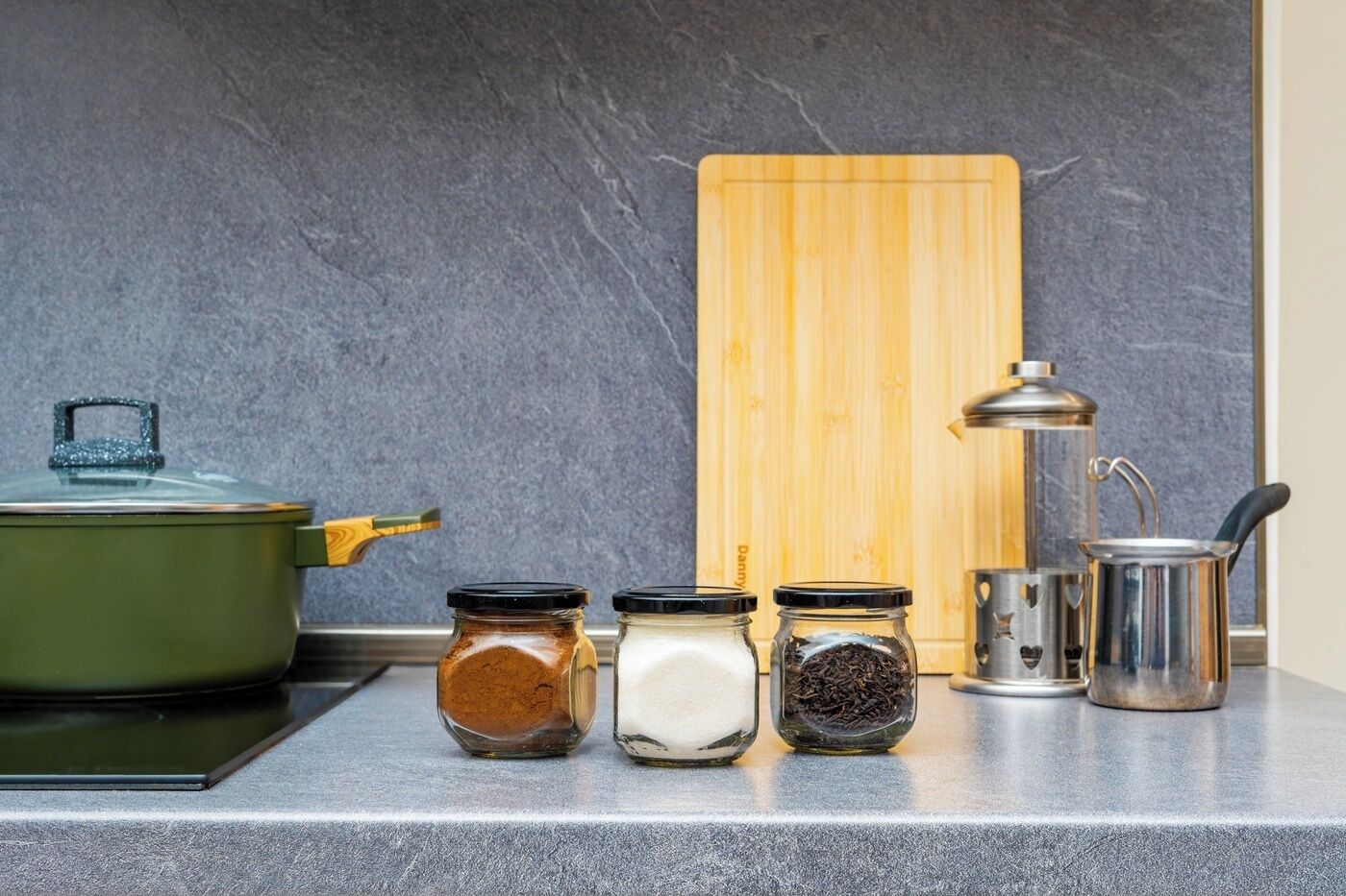 Un piano di lavoro in cucina con una pentola verde, tre barattoli di vetro con diversi contenuti, un tagliere di legno e un bollitore metallico.