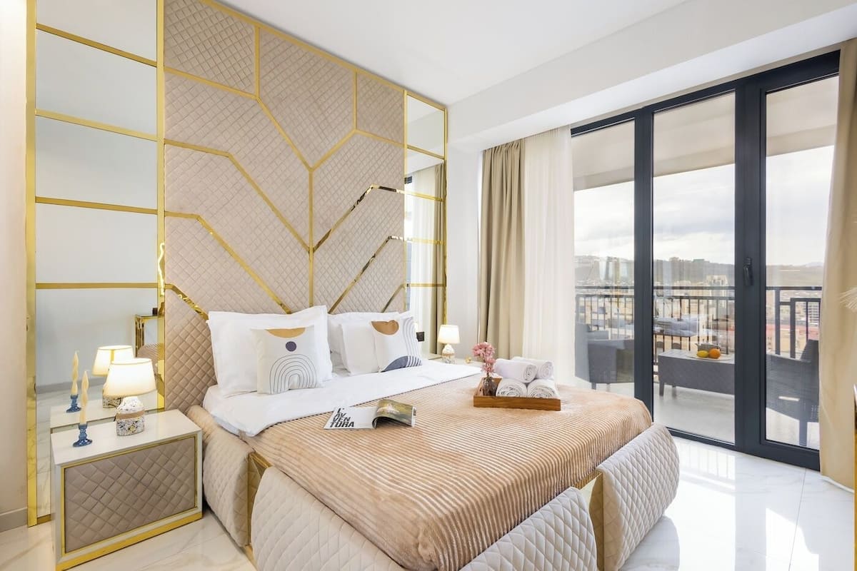 A modern hotel room with a large bed, a bedside table with a lamp, a view of the city through large windows, and a balcony with a table and chairs.