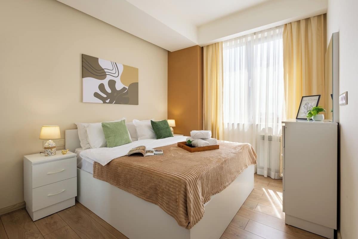A hotel room with a bed, bedside tables, a lamp, a framed picture, a plant, and a radiator.
