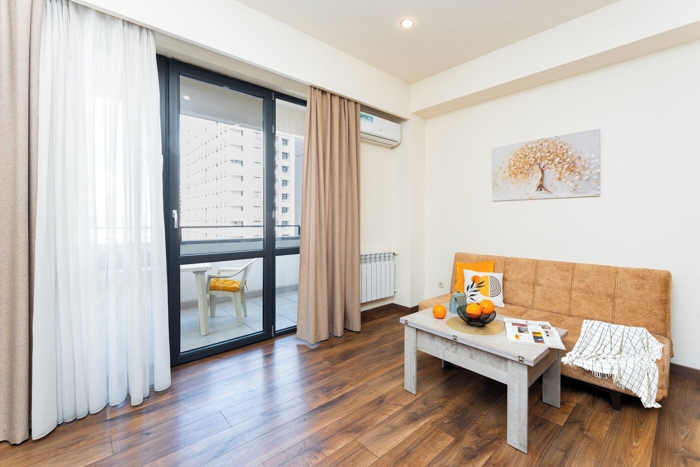 A modern living room with a wooden floor, a sofa, a coffee table with fruits, and a view of a cityscape through a glass door.