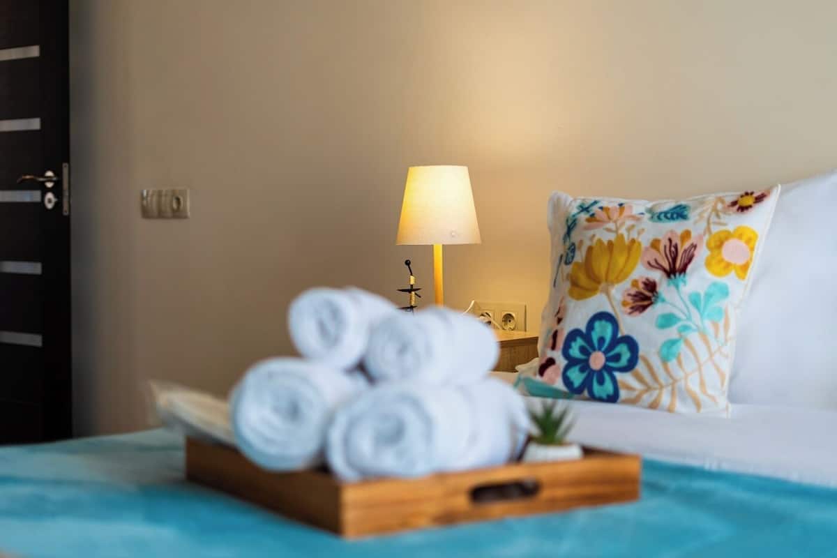 A neatly made bed with a floral pillow, a bedside lamp, and a tray of rolled towels.
