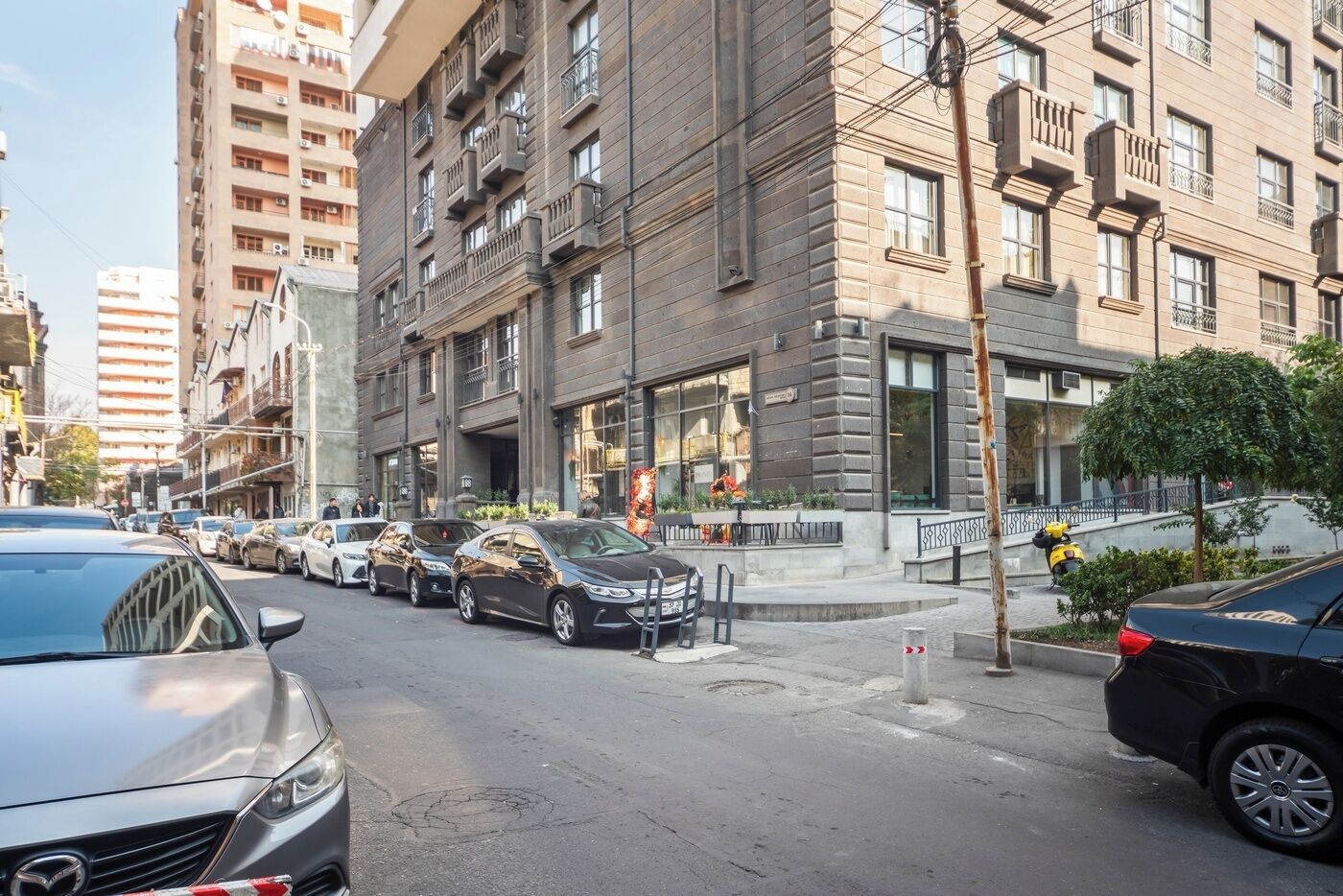 Vista de la calle con una fila de coches aparcados, un edificio con balcones y un paso de peatones.