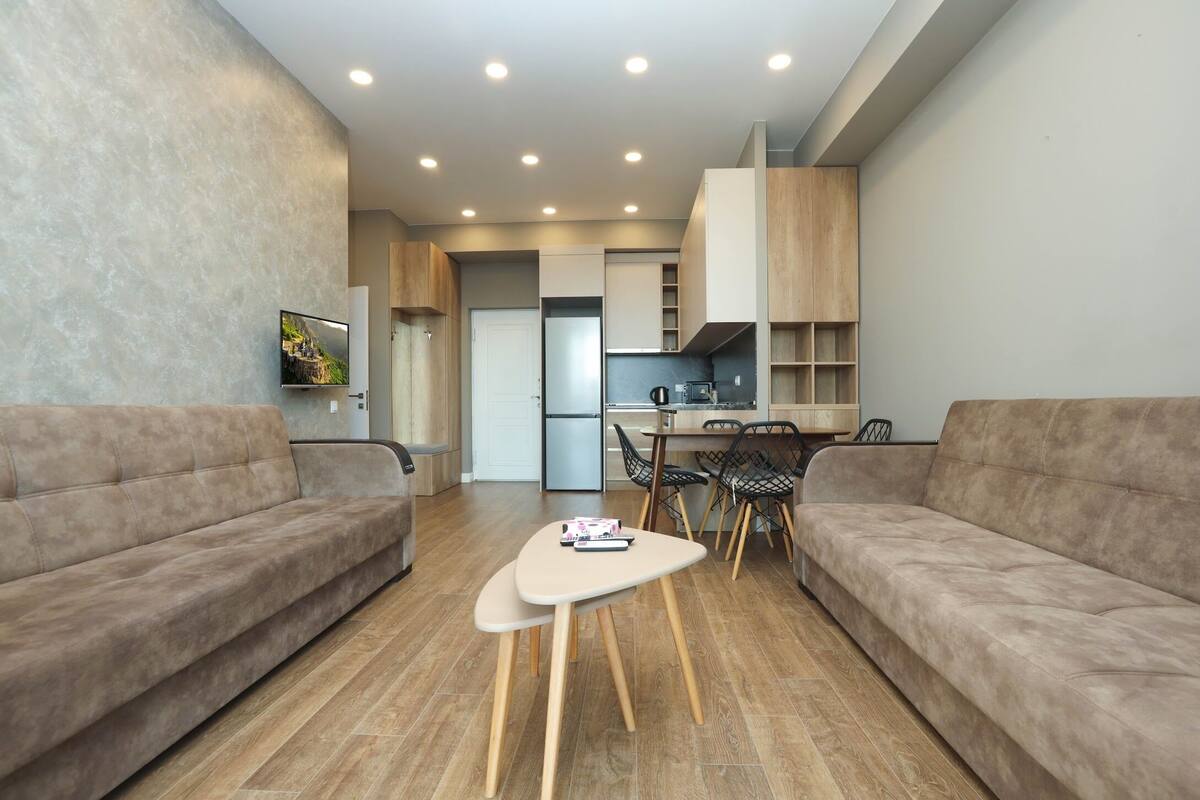 A modern living room with a sofa, a coffee table, and a TV. There is a kitchen area with cabinets and a refrigerator.