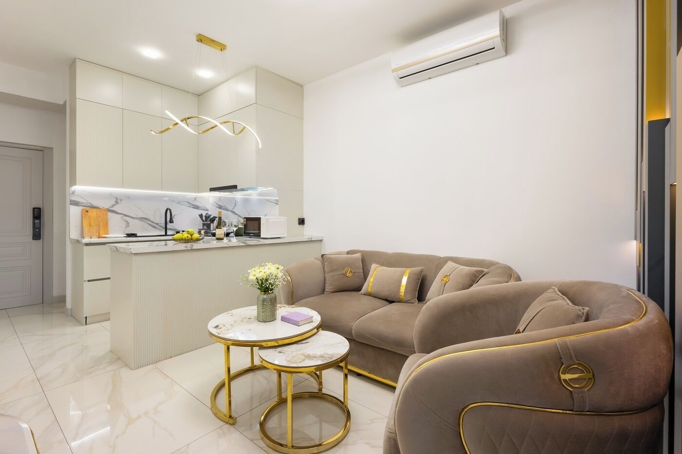 Un salon moderne avec un canapé beige, une table basse en marbre et un coin cuisine doté de meubles blancs et d’un four encastré.