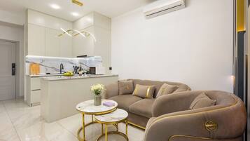 Un salon moderne avec un canapé beige, une table basse en marbre et un coin cuisine doté de meubles blancs et d’un four encastré.