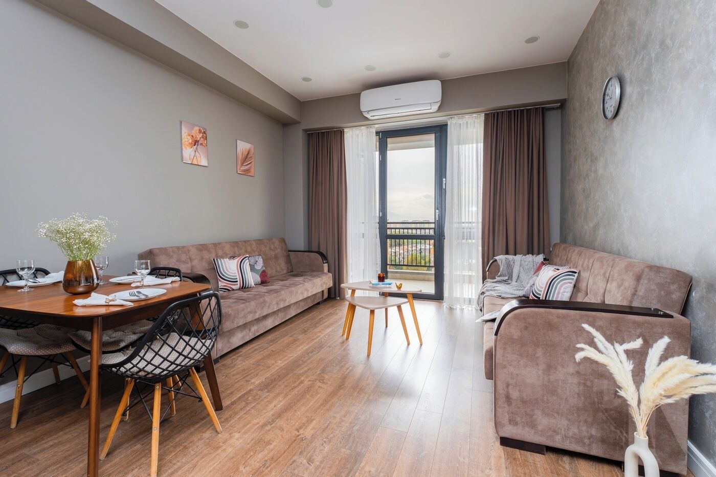 A modern living room with a sofa, a dining table with chairs, and a small coffee table. There is a balcony with a view, and an air conditioning unit on the wall.