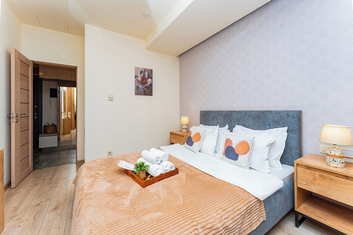 A hotel room with a bed, wooden headboard, bedside tables, a nightstand, and a wall-mounted lamp.