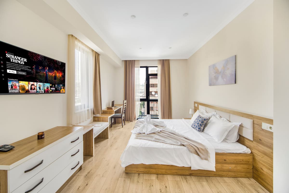 A hotel room with a bed, a desk, a chair, a TV mounted on the wall, and a window with curtains.