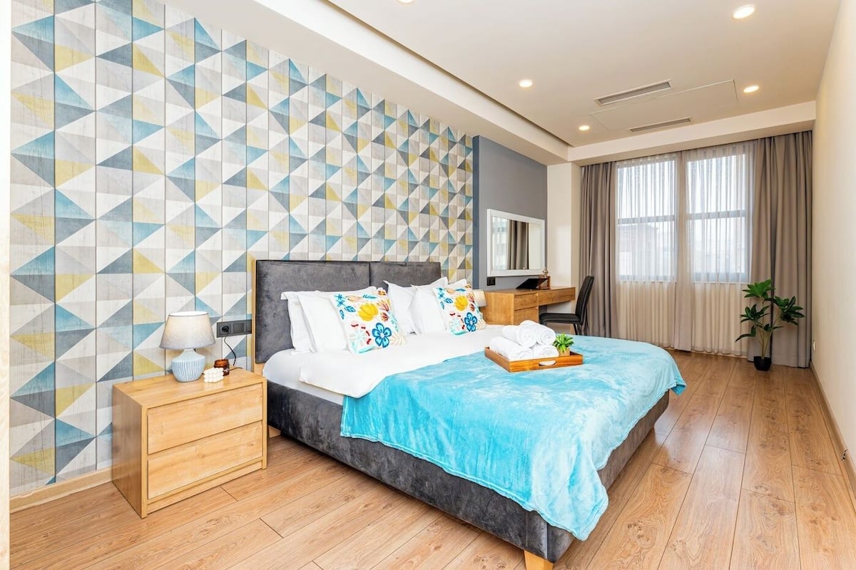 A modern hotel room with a geometric-patterned wall, a large bed with a blue blanket, wooden nightstands, a desk, and a potted plant.