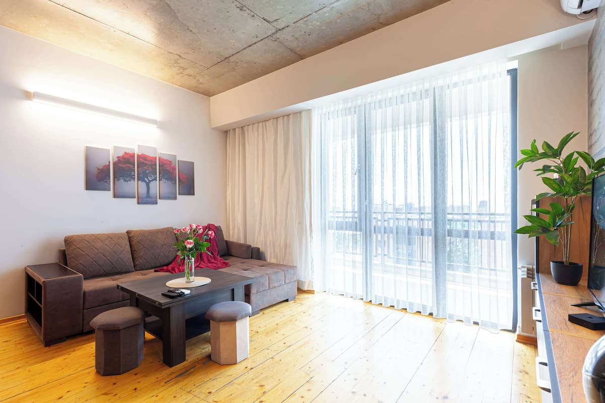 A modern living room with a sofa, coffee table, and ottomans, large windows with sheer curtains, and a potted plant.
