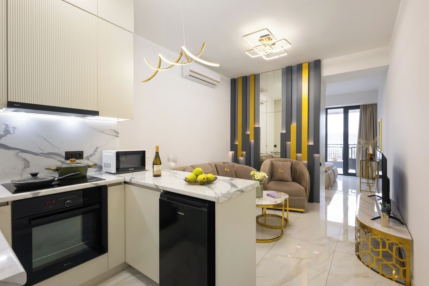 A modern kitchen with a microwave, stove, and dishwasher. There is a dining area with a sofa and a small table. The room has a large window and a decorative wall panel.