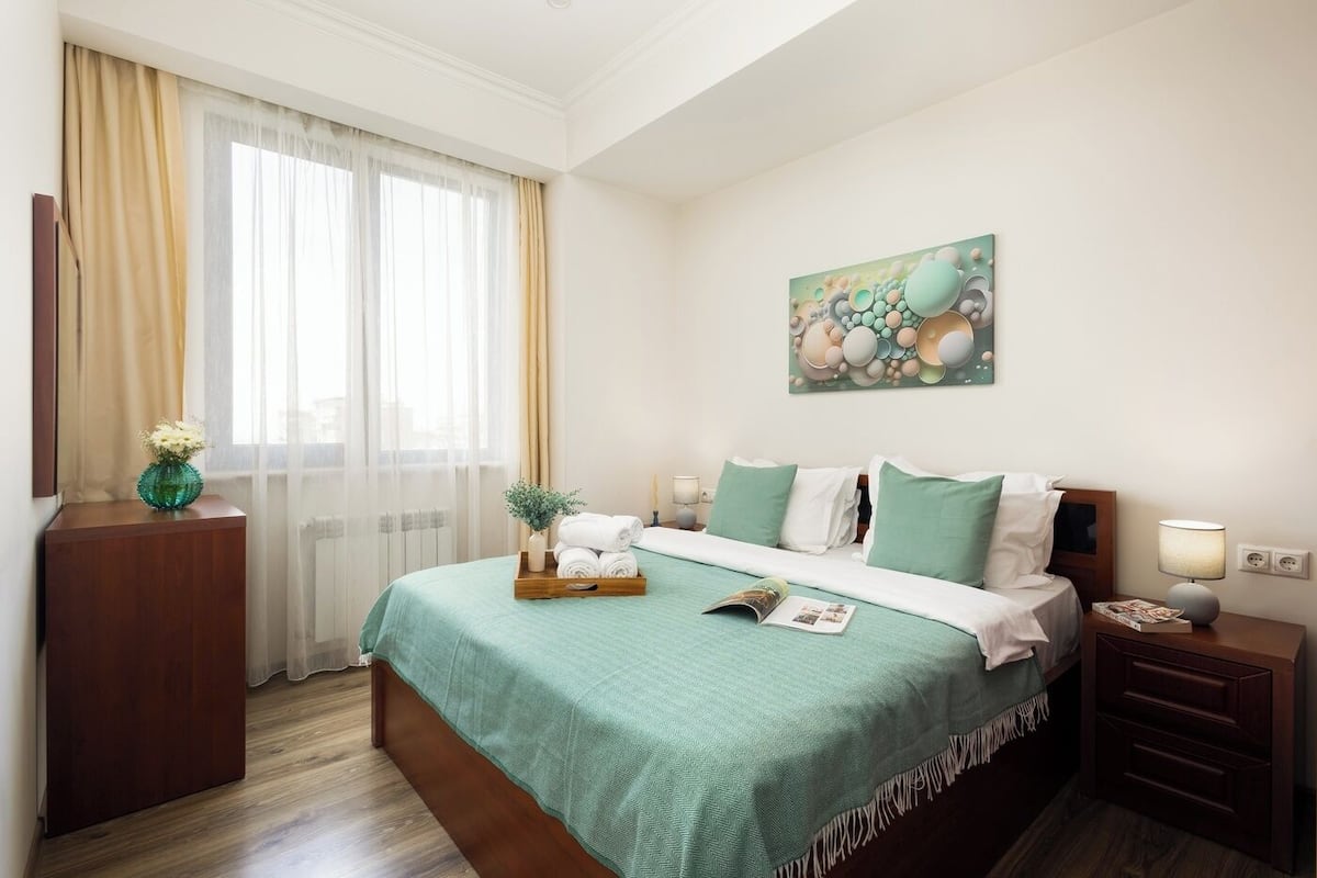 A hotel room with a bed, a bedside table with a lamp, a nightstand, a window with curtains, and a wall-mounted artwork.