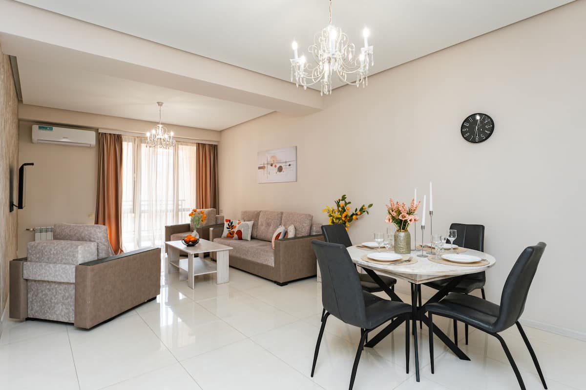 A modern living room with a dining area, a sofa set, a chandelier, and a clock on the wall.