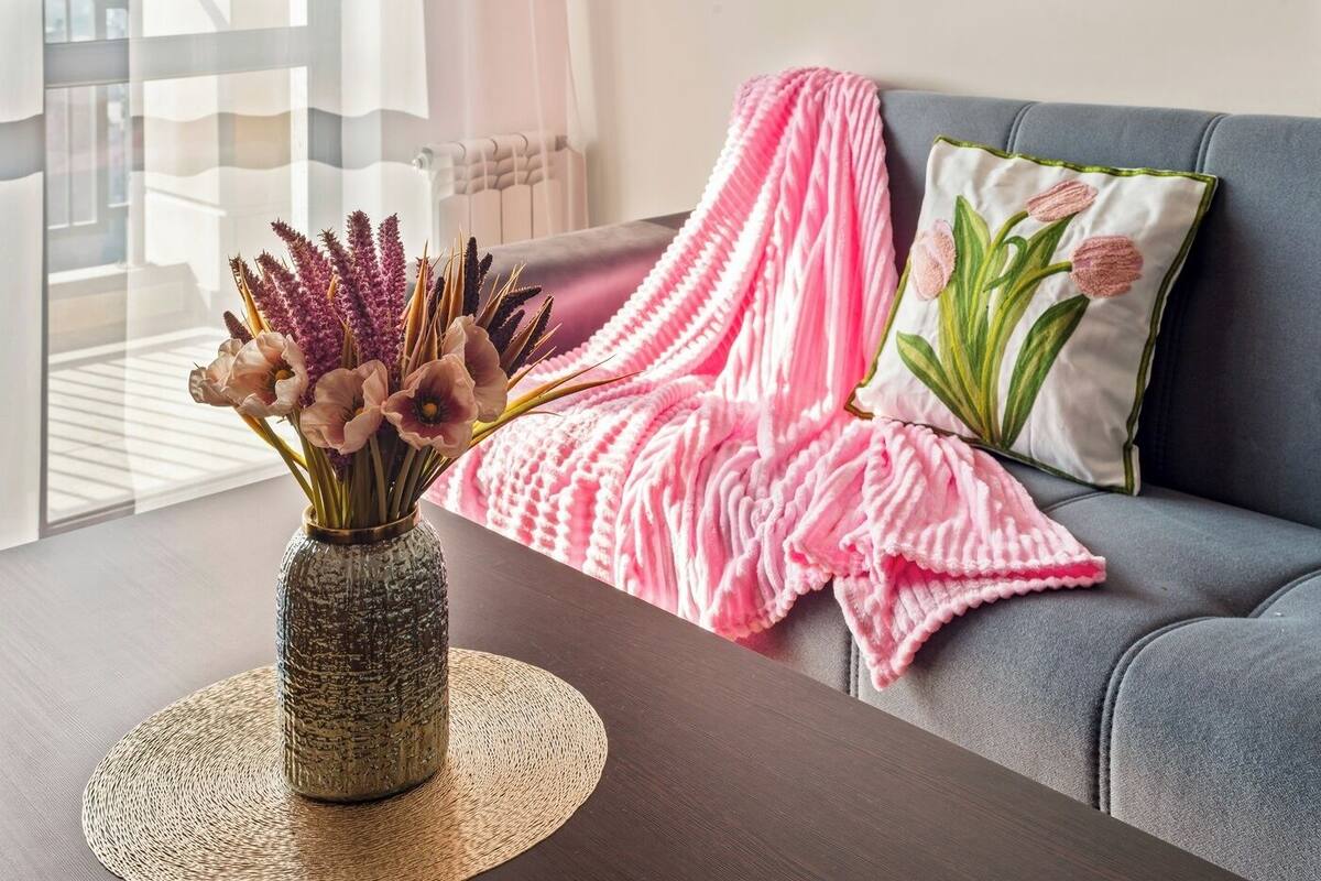 A living room with a grey sofa, a pink blanket, a decorative pillow with a floral design, and a vase with flowers on a round woven placemat.