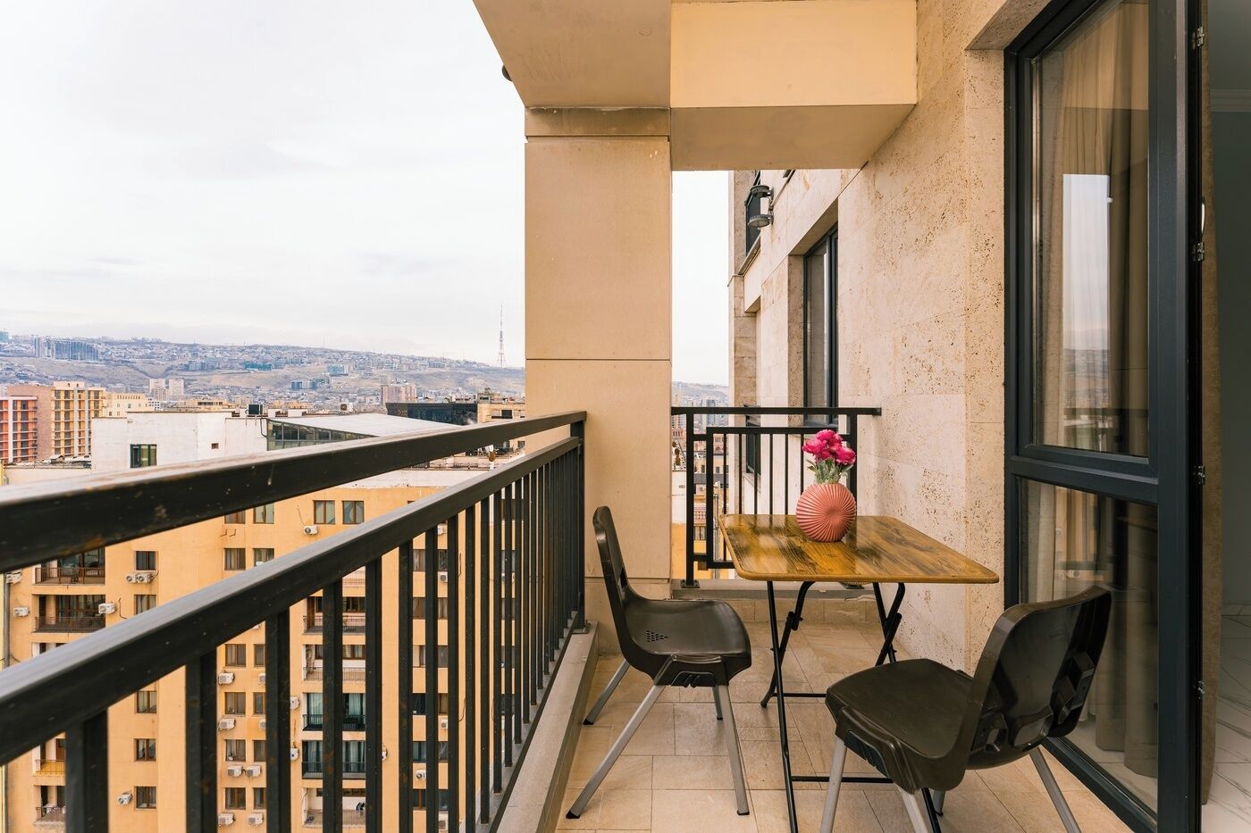 A balcony with a table and chairs, a vase with flowers, and a city view.
