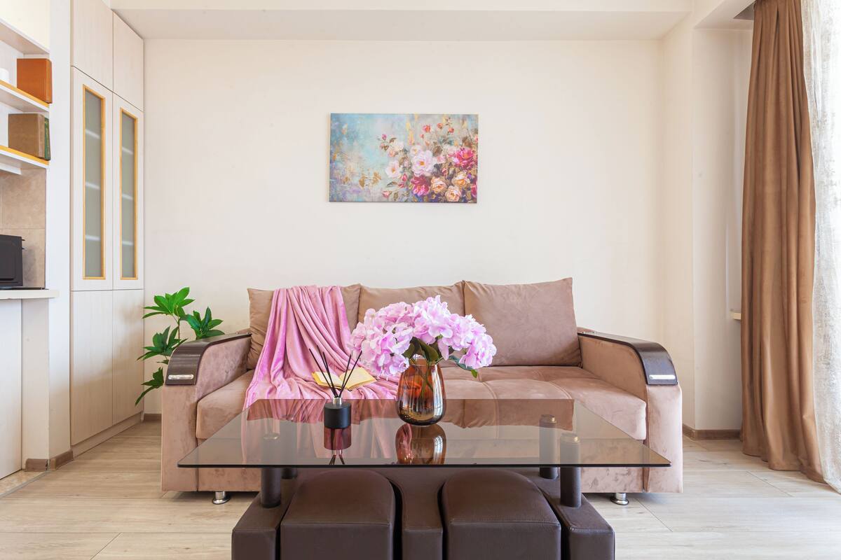 A modern living room with a glass coffee table, a beige sofa, a pink throw, and a floral painting on the wall.