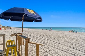 Une plage avec un parasol bleu portant l’inscription « Sandestin », des chaises en bois et un tabouret jaune.