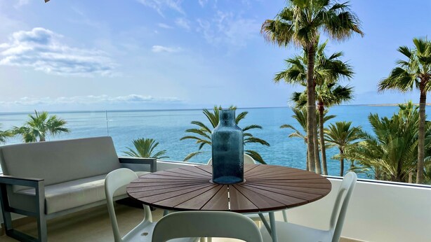 A balcony with a round wooden table, two white chairs, a blue vase, and a view of the sea with palm trees. - Relaxing Ocean Views, Closed to the Beach (San Bartolomé de Tirajana)
