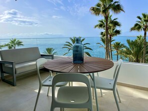 Un balcon avec une table ronde en bois, deux chaises blanches, un vase bleu et une vue sur la mer, avec des palmiers.