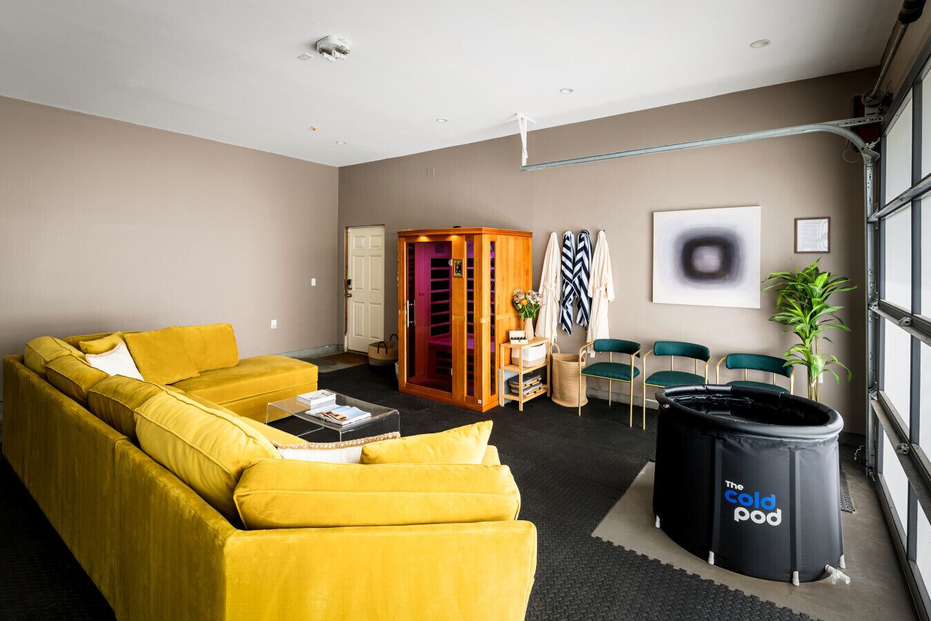 A modern living room with a yellow sofa, a small coffee table, a trash bin labeled "The Cold Pod," and a view of the outside through large windows.