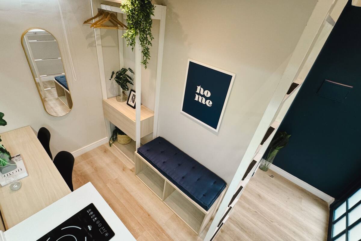 Une pièce moderne avec un plancher en bois, un banc, un miroir, une plante suspendue et un panneau indiquant « chez soi » sur le mur.
