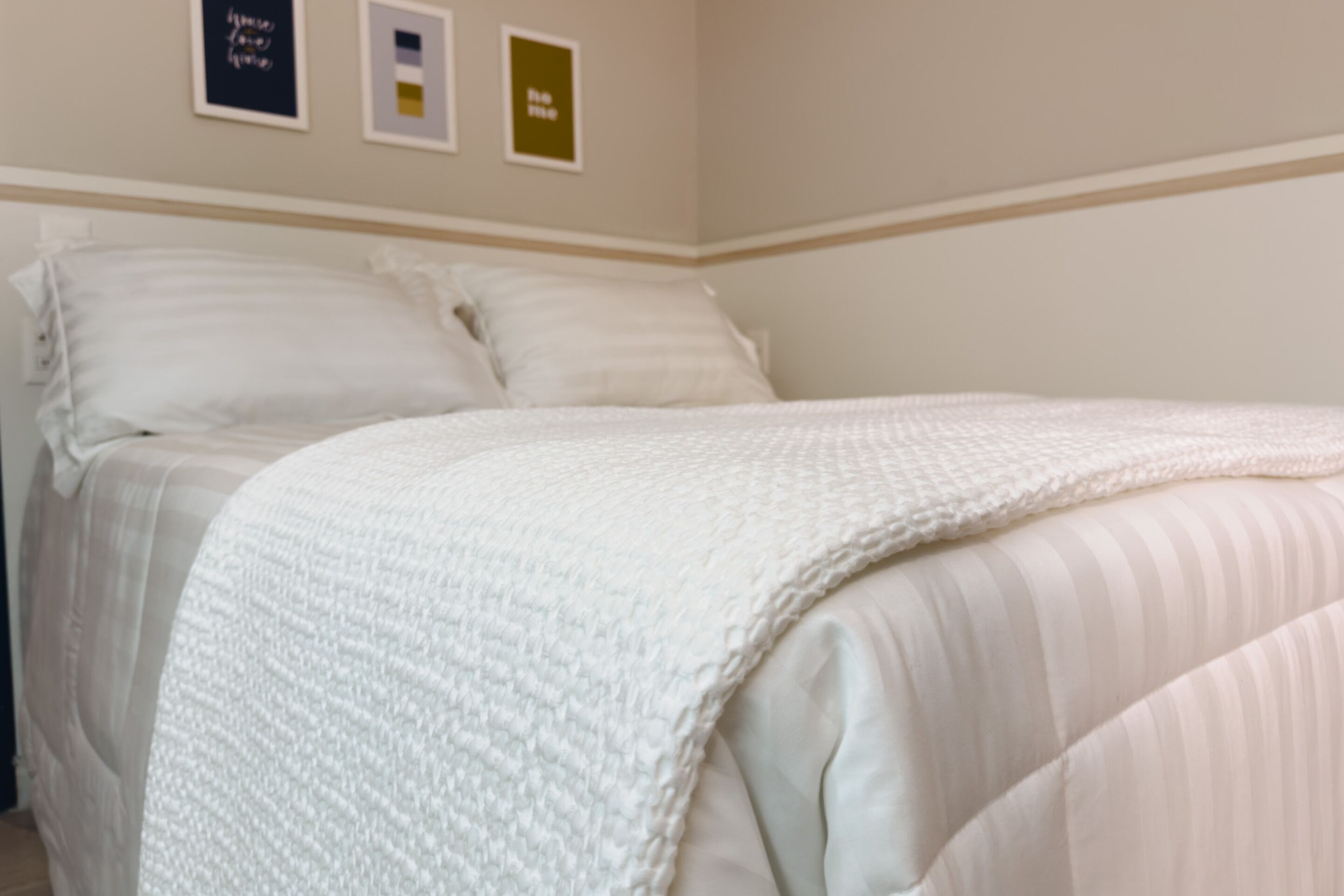 Un lit bien fait, avec du linge de lit blanc et des oreillers, une couverture blanche à texture intéressante et un mur orné de trois tableaux encadrés.