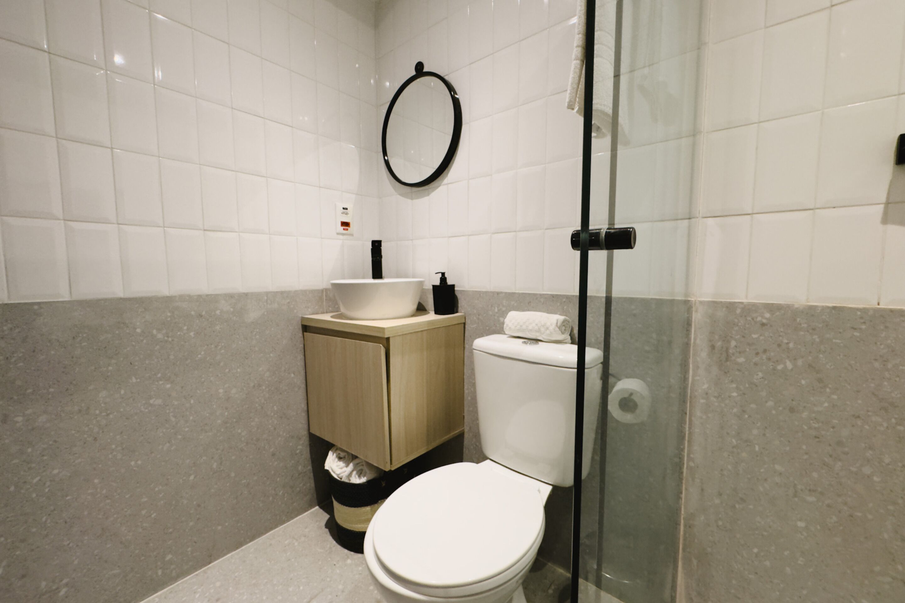 A modern bathroom with a white toilet, a sink with a beige cabinet, a glass shower enclosure, and a round mirror on the wall.