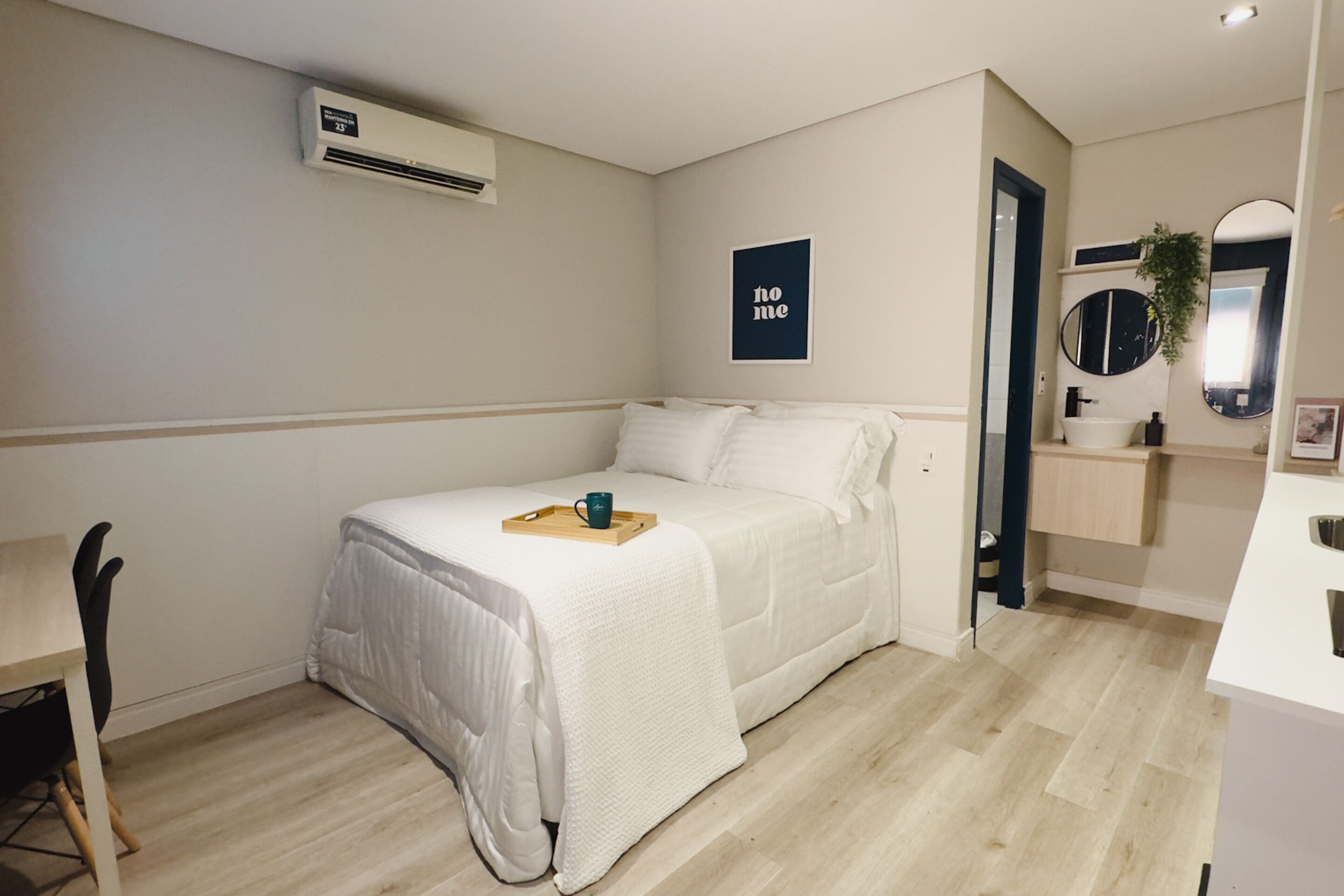 A bedroom with a bed, a bedside table, a chair, a bedside plant, and a bathroom with a sink and mirror.