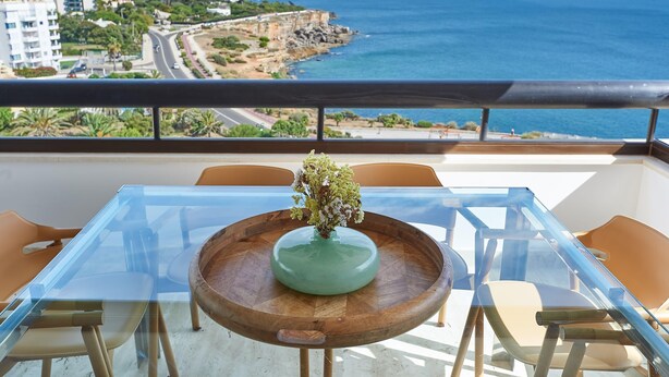 A balcony with a glass table, wooden chairs, and a vase with flowers. A view of the ocean and buildings is visible. - Luxury Ocean View Apartment by Portugal Portfolio (Cascais)