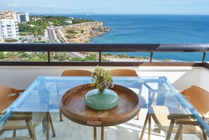 A balcony with a glass table, wooden chairs, and a vase with flowers. A view of the ocean and buildings is visible. - Luxury Ocean View Apartment by Portugal Portfolio (Cascais)