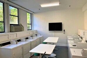 A classroom with white desks, a flat-screen TV mounted on the wall, and windows with blinds. - SpecialOfferPool/StepsToBeach/CharmStudio (Palm Beach)