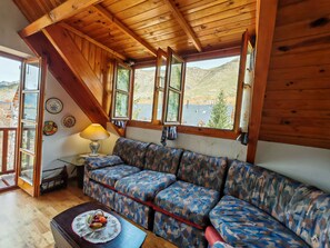 A cozy room with a wooden ceiling, a patterned sofa, a table with fruit, and a window with a view of mountains.