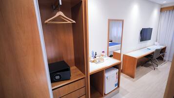 Habitación de hotel con armario de madera, escritorio, silla, espejo, cama y un refrigerador pequeño.