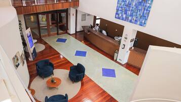 A spacious lobby with a wooden floor, blue armchairs, a reception desk, and a staircase.