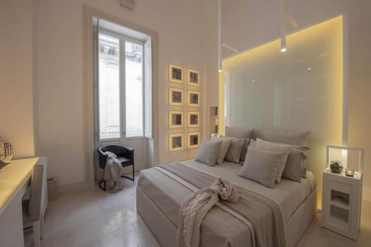 A modern bedroom with a large bed, a desk, and a chair. There are framed pictures on the wall and a window with blinds.
