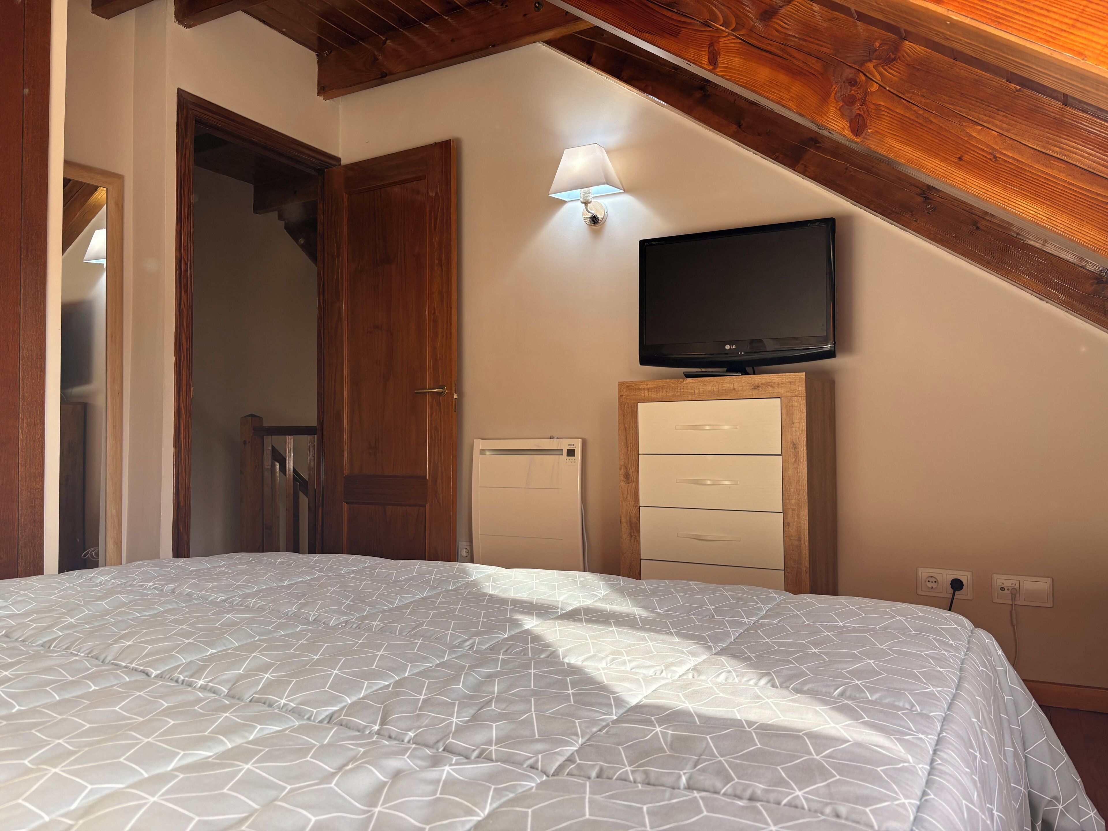 A bedroom with a bed, a TV mounted on the wall, a wooden cabinet, and a ceiling with exposed beams.