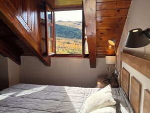 Une chambre avec un plafond en pente en bois, un lit recouvert d’une courtepointe matelassée, une table de chevet avec une lampe et une fenêtre donnant sur un paysage vallonné.