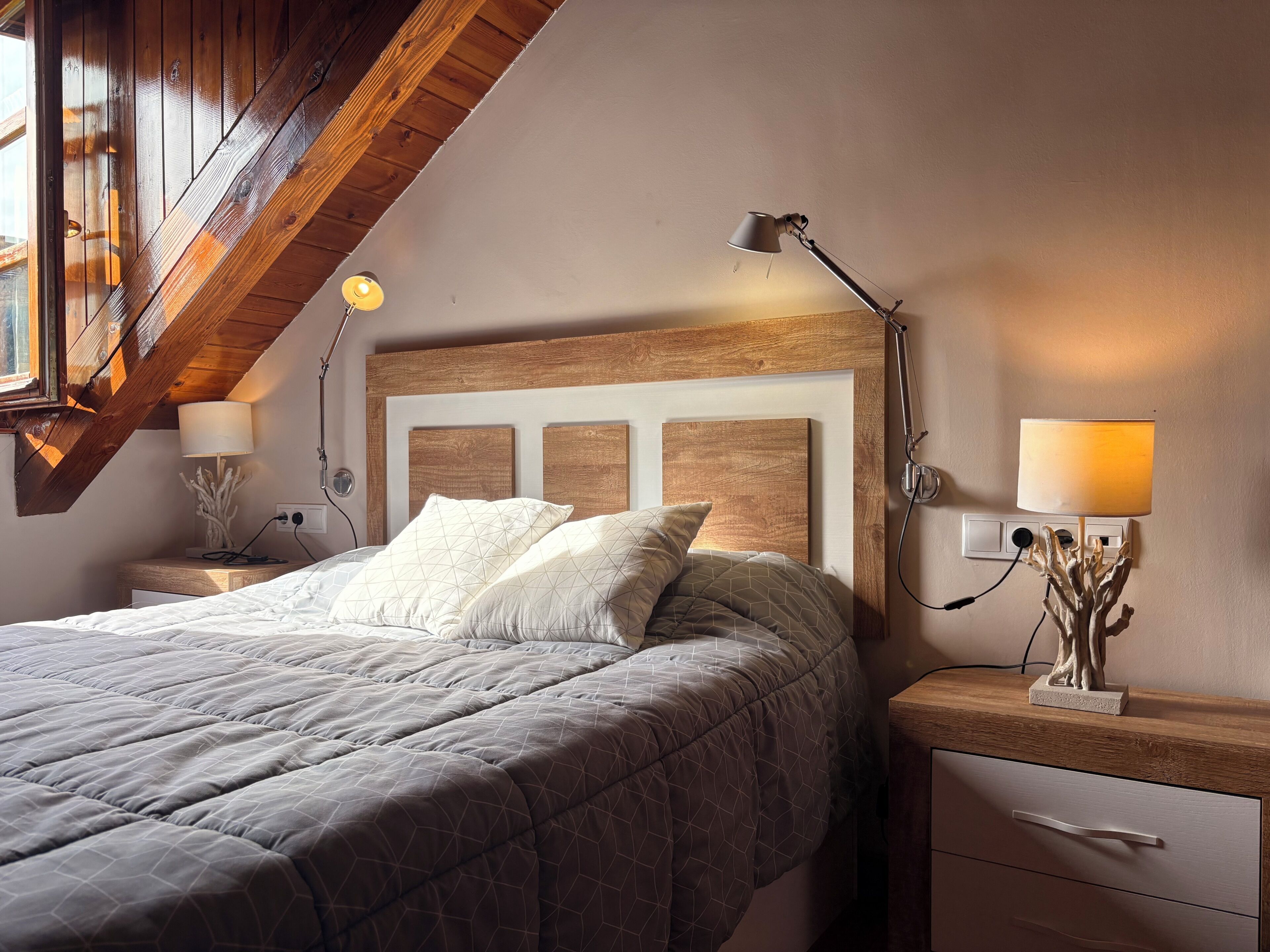 A bedroom with a wooden headboard, a bed with a geometric-patterned duvet, bedside lamps, and a nightstand with a decorative item.