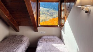 A room with two beds, a window with a view of autumn foliage, and a wall-mounted lamp.