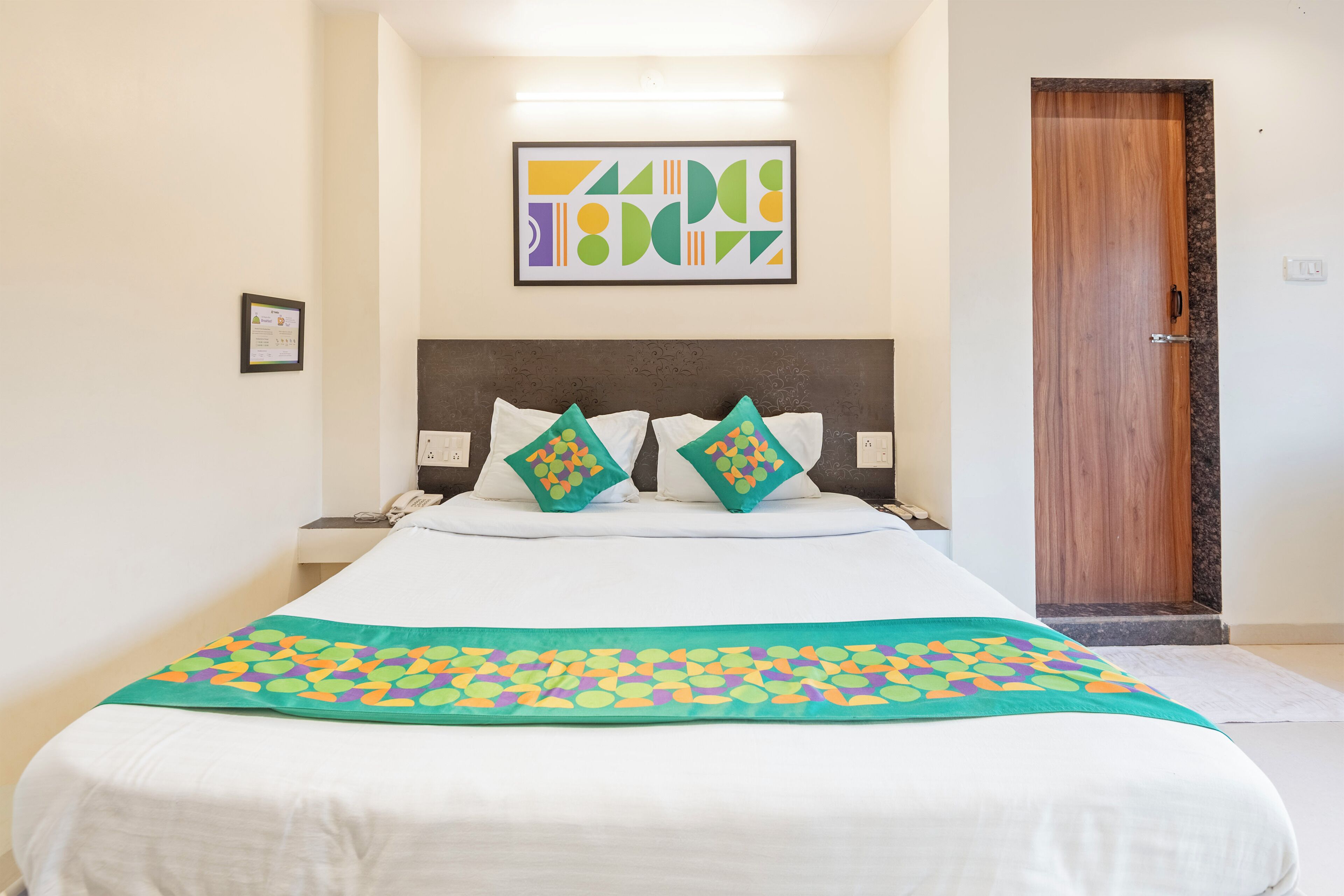 A hotel room with a bed, a headboard, a wall-mounted artwork, a door, and a bedside table with a phone.