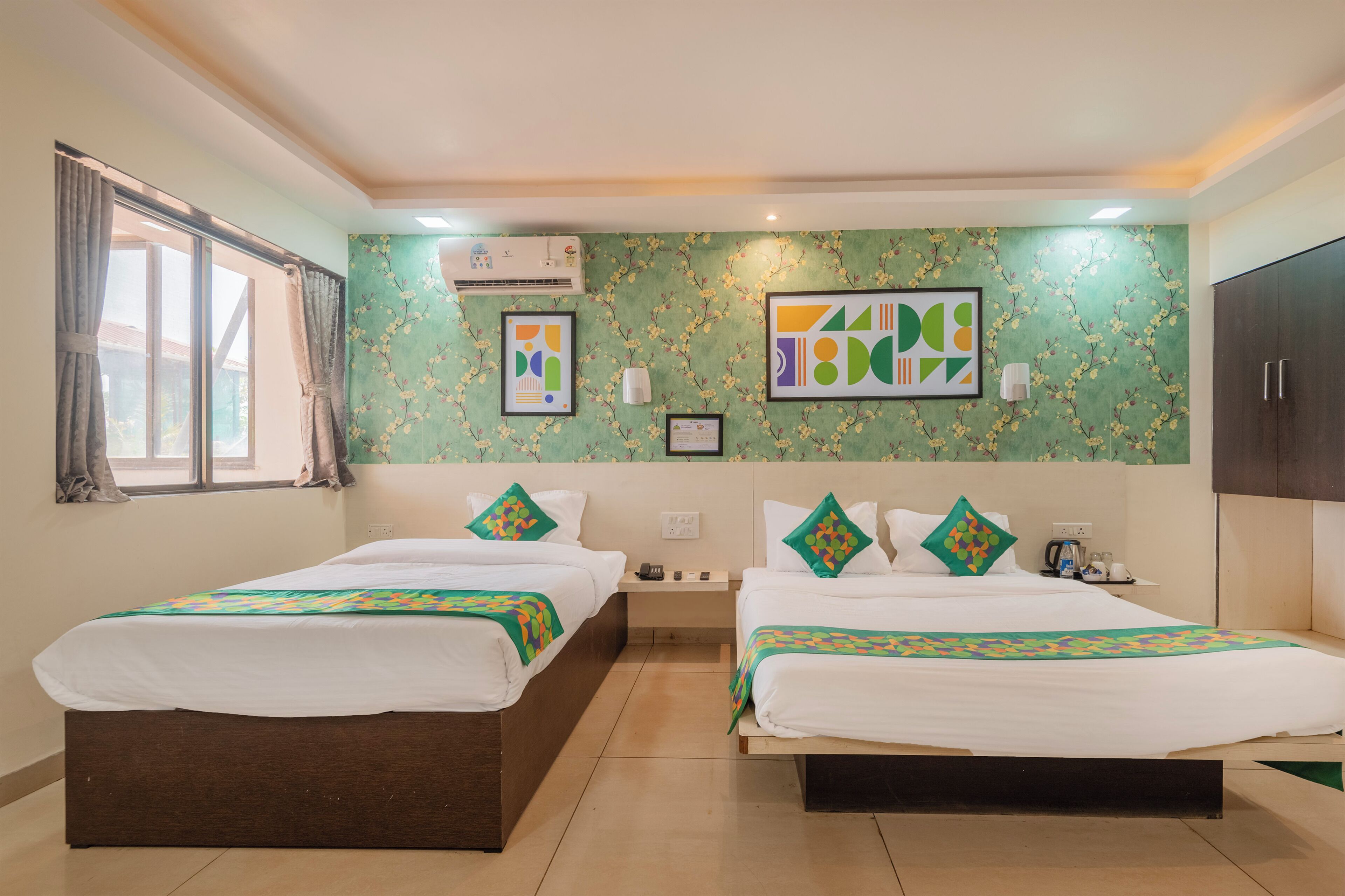 A hotel room with two beds, a window with curtains, an air conditioning unit, and a wall with floral wallpaper and framed artwork.