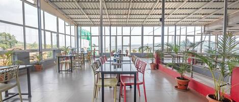 A spacious indoor area with a long table and chairs, large windows, and potted plants.