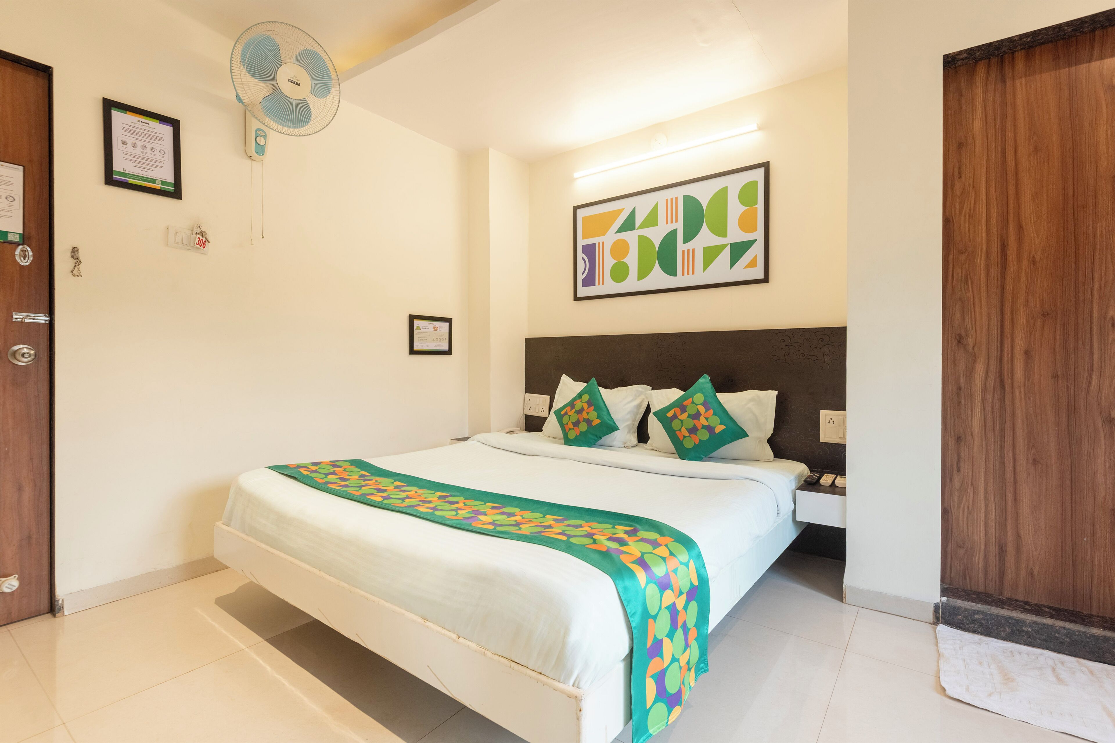 A hotel room with a bed, a wall-mounted fan, a framed certificate, and a painting.