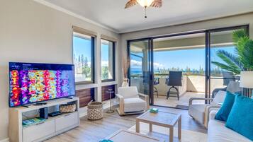A modern living room with a television, sofa, and coffee table, a balcony with chairs, and a view of the ocean.