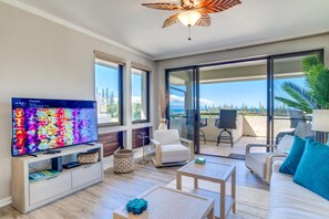 A modern living room with a television, sofa, and coffee table, a balcony with chairs, and a view of the ocean.