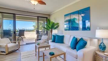 A modern living room with a white sofa, blue cushions, a wooden coffee table, and a ceiling fan. There is a large window with a view of the outdoors.