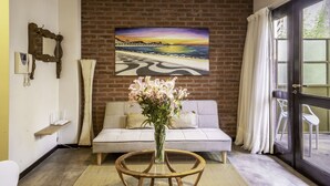 A living room with a sofa, a round coffee table with a vase of flowers, a painting of a beach scene, and a brick wall. - 32-rustico Natural.2951 José (Ciudad Autónoma de Buenos Aires, Recoleta)