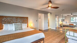 A hotel room with a large bed, a ceiling fan, a wooden headboard, a bedside lamp, and a view of another room through a glass door.