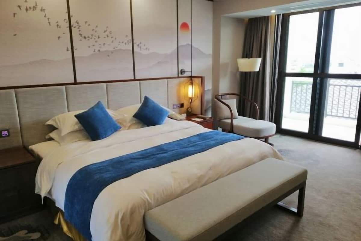 A hotel room with a large bed, a bench, a chair, a wall-mounted lamp, and a view of the outdoors through a large window.