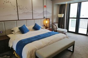 A hotel room with a large bed, a bench, a chair, a wall-mounted lamp, and a view of the outdoors through a large window.